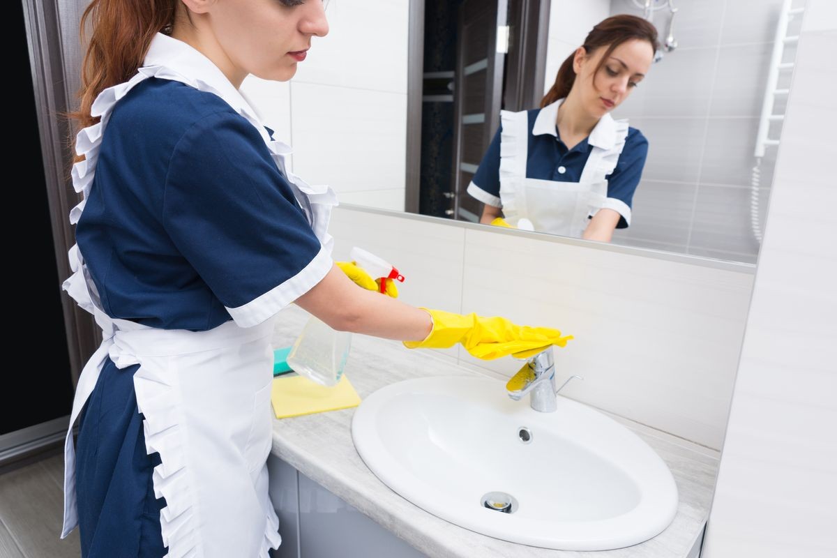 Housekeeper in a neat uniform and apron servicing a hotel bathroom washing and cleaning the hand basin and faucet with a yellow cloth, reflected in the mirror